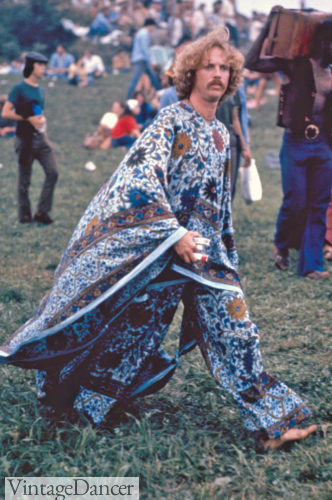 A hippie man in robes walks through grass. Behind him are multiple men in more typical hippie clothing, such as bright pants and simple shirts.