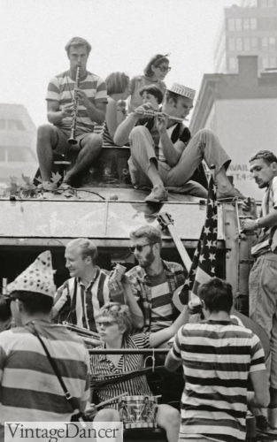 The Merry Pranksters "tootling" with various instruments aboard Furthur in 1964. Many are wearing stripes or basic clothing, with the occasional odd accessory such as a birthday hat or a large American flag.